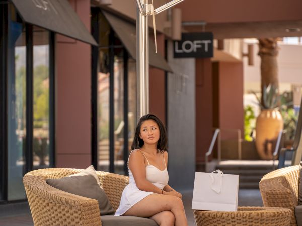 A person sits on a wicker chair under an umbrella at an outdoor mall with shopping bags nearby.