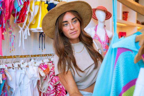 A woman wearing a hat is shopping for clothes in a store with colorful swimsuits displayed on racks.