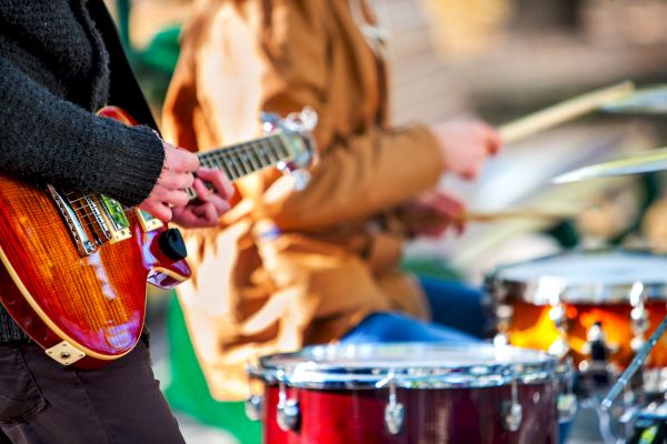 The image shows people playing musical instruments, specifically a guitar and drums, in an outdoor setting.