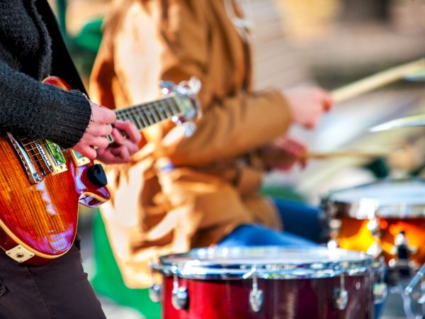 The image shows people playing musical instruments, specifically a guitar and drums, in an outdoor setting.