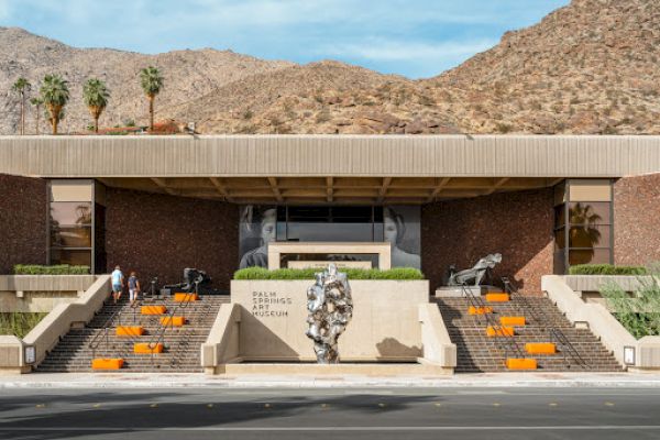 The image shows the exterior of the Palm Springs Art Museum, with steps, sculptures, and a desert mountain backdrop under a blue sky.