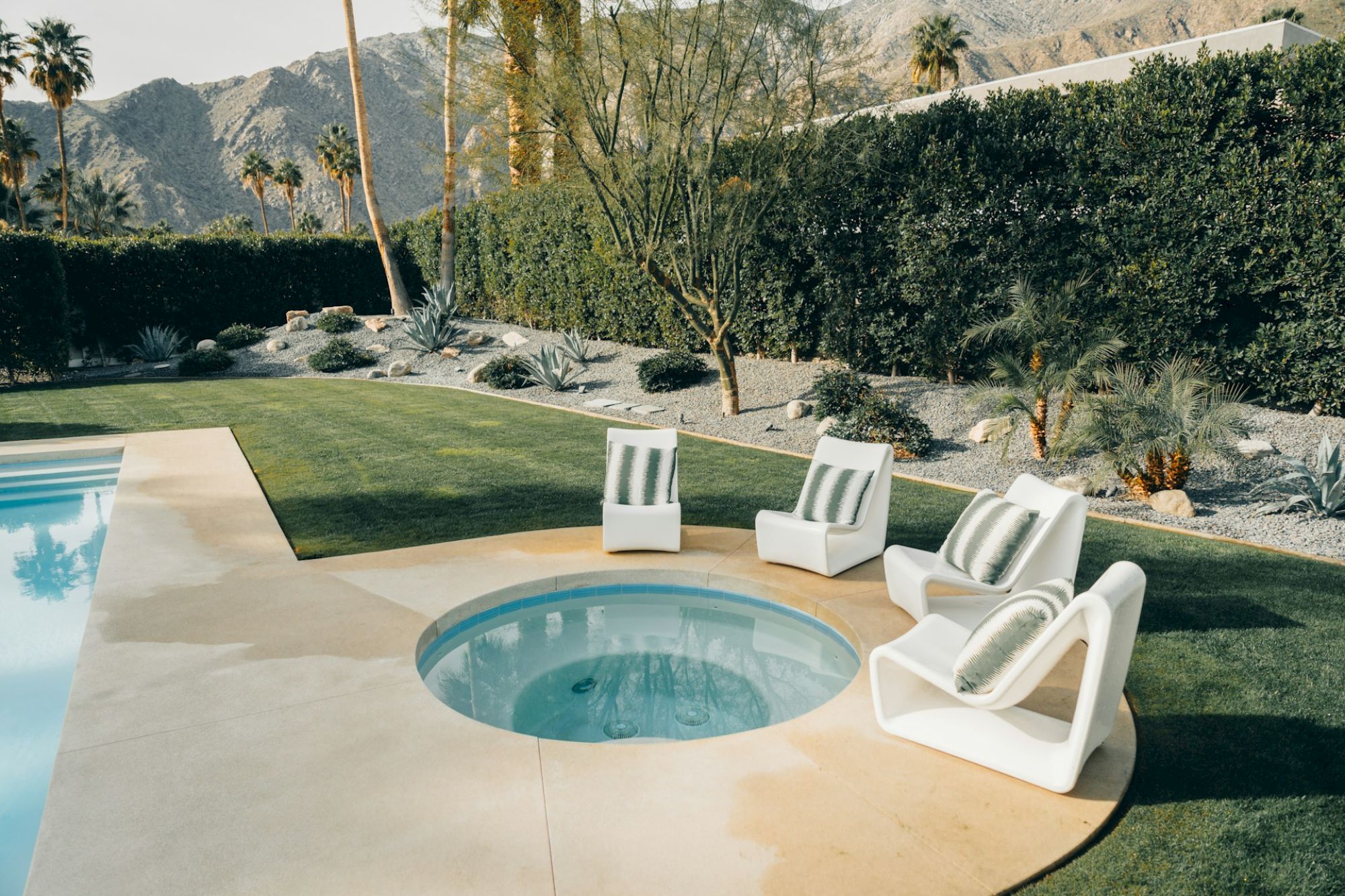 A backyard with a pool and hot tub, surrounded by lounge chairs and greenery, set against a backdrop of mountains and tall trees.