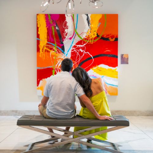 A couple sits on a bench, facing a vibrant abstract painting with bold reds and oranges in a gallery setting.