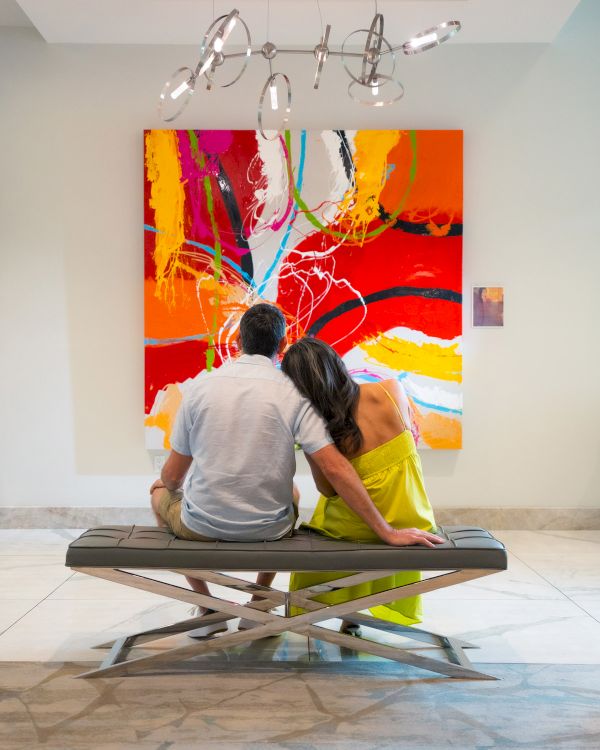 A couple sits on a bench, facing a vibrant abstract painting with bold reds and oranges in a gallery setting.