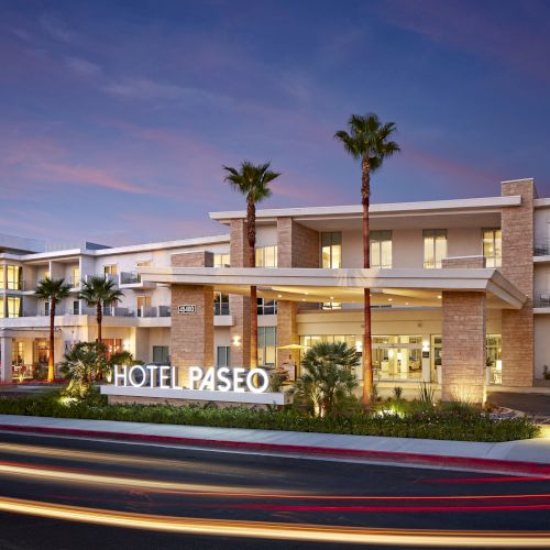 A modern hotel exterior with a lit "HOTEL PASEO" sign, palm trees, and light trails from passing vehicles under a dusk sky.