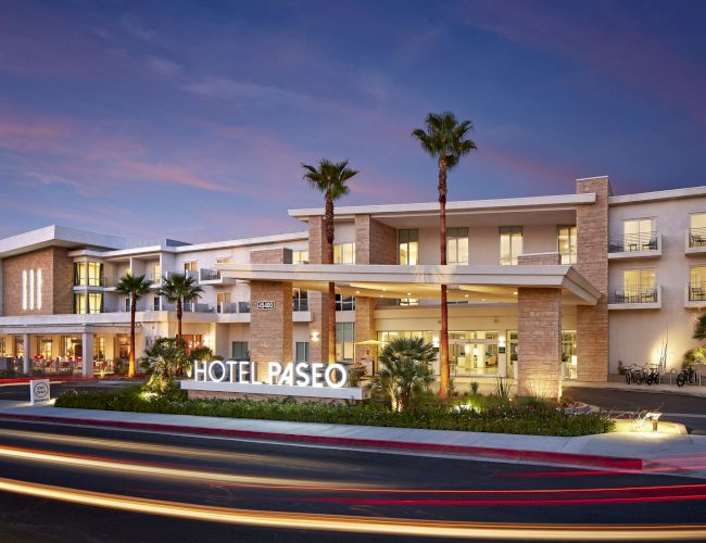 A modern hotel exterior with a lit "HOTEL PASEO" sign, palm trees, and light trails from passing vehicles under a dusk sky.