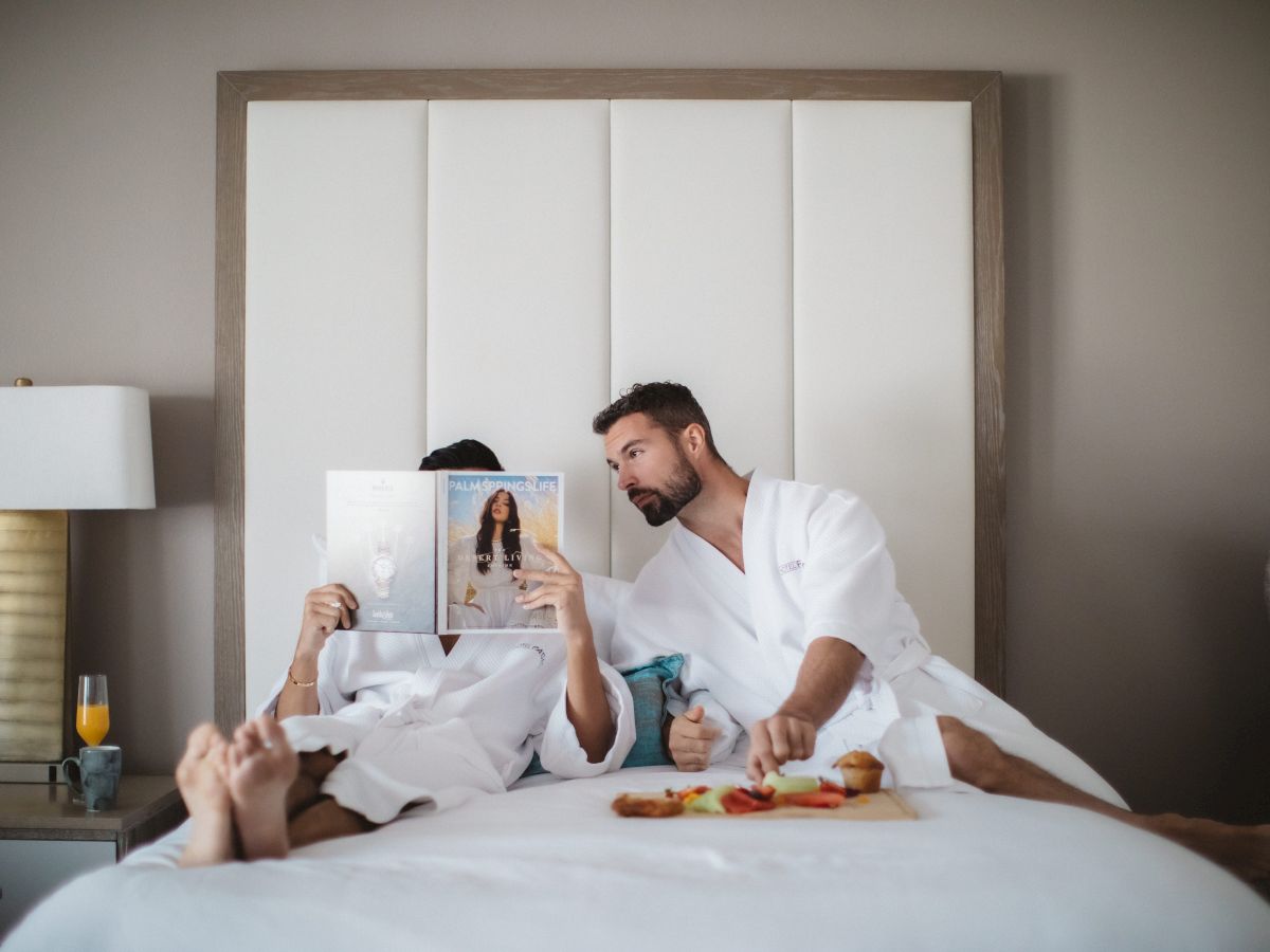 Two people in bathrobes are on a bed; one reads a magazine, the other reaches for food on a tray. A bedside lamp and drink are visible.