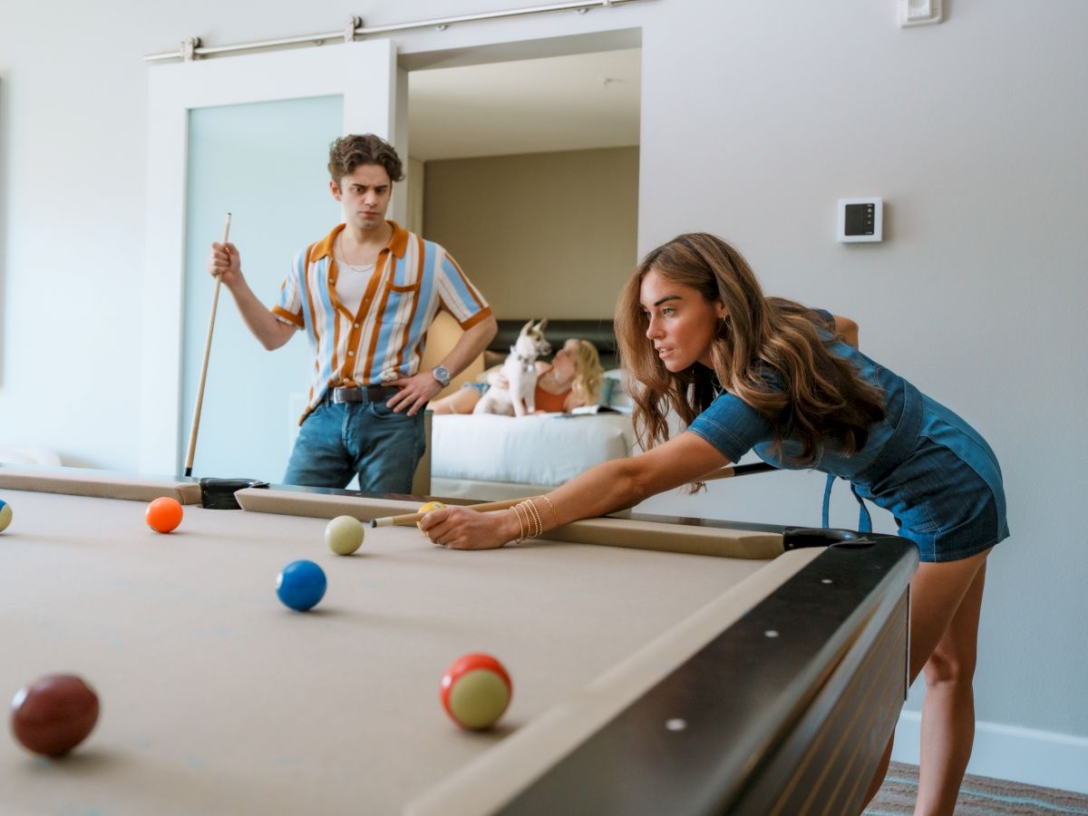 Two people are playing pool in a room with a dog on a bed in the background.