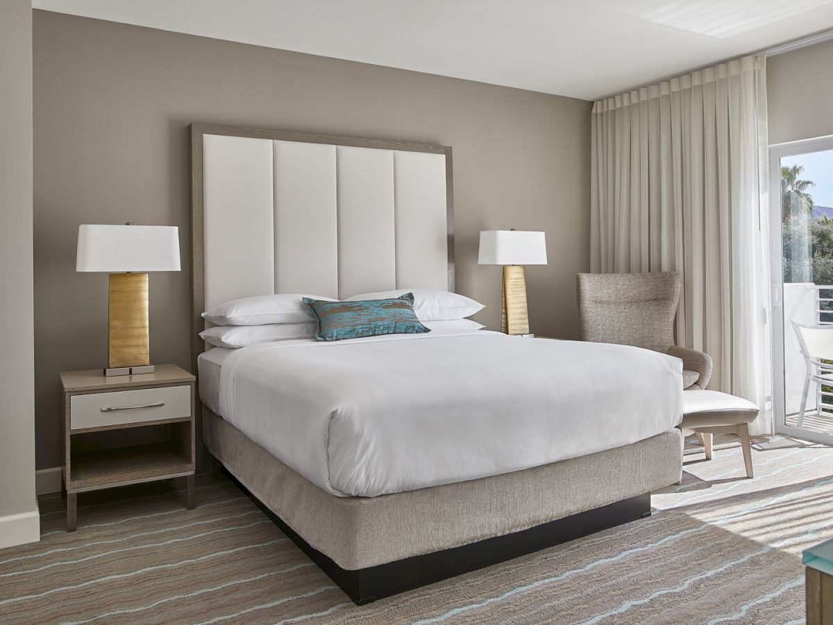 A modern bedroom with a large bed, two bedside tables with lamps, a chair, and a window with curtains. Neutral tones create a calming ambiance.