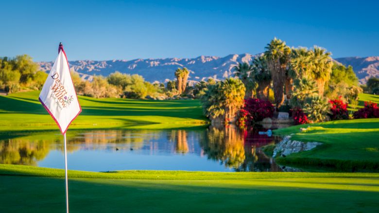 A golf course with a flag labeled "Desert Willow Golf Resort," surrounded by palm trees, a pond, and mountains in the background.