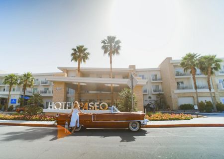 A person stands beside a vintage car in front of Hotel Paseo, surrounded by palm trees under a bright sky.