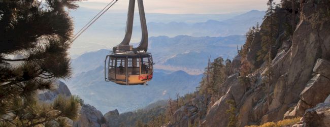 A tramway car suspended above rocky mountains with pine trees, against a vast landscape and hazy sky in the background.