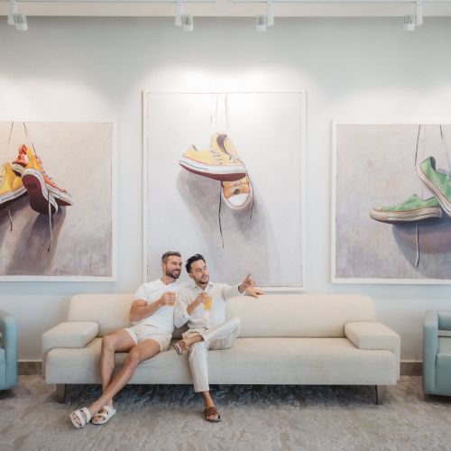 Two people sitting on a sofa under paintings of sneakers. There are two blue armchairs flanking the sofa in a modern, bright room.