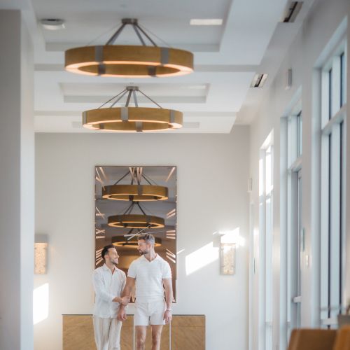 A couple walks down a modern hallway with circular chandeliers and large windows.