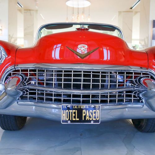 A vintage red car with a "HOTEL PASEO" California license plate, displayed indoors with a shiny front grille and headlights.