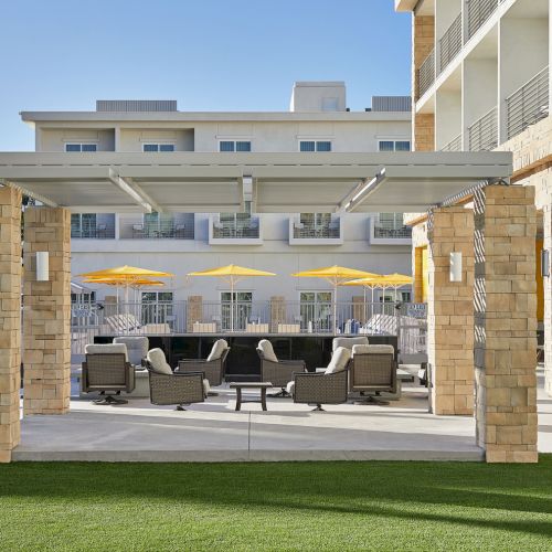 The image shows an outdoor seating area with chairs, tables, and yellow umbrellas under a pergola, with a modern building in the background.