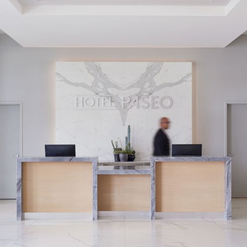 The image shows a hotel reception area with two desks and a wall sign reading "HOTEL PASEO." A person is walking behind the desks.