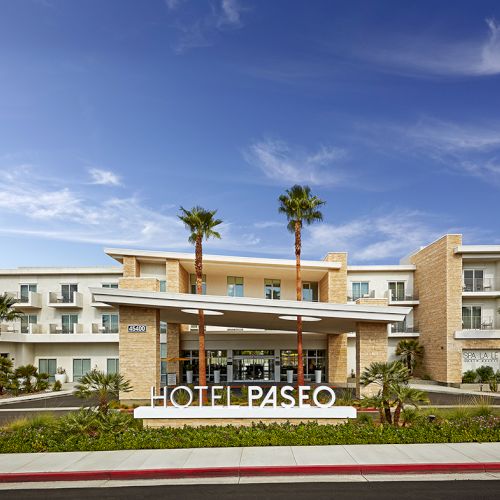 The image shows the exterior of Hotel Paseo, featuring palm trees and a modern entrance under a clear blue sky.