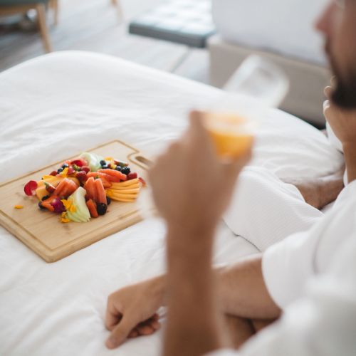 A person in a white robe is sitting on a bed with a wooden board of assorted fruits, holding a glass of orange juice.