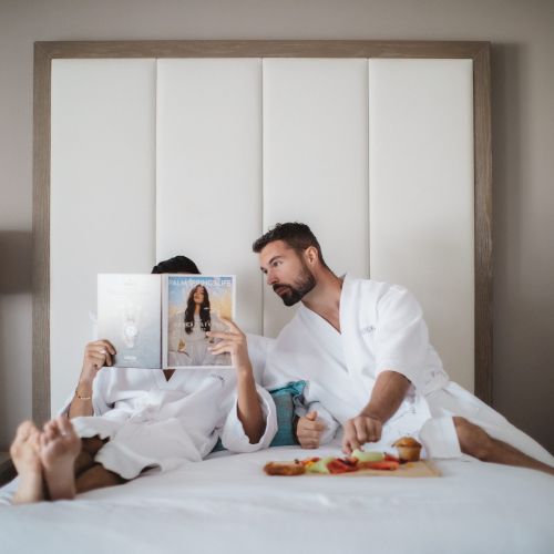 Two people in bathrobes relax on a bed, one reads a magazine while the other reaches for food on a tray.