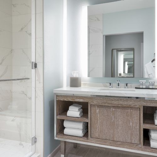 The image shows a modern bathroom with a glass shower, illuminated mirror, and a vanity with shelves holding neatly folded towels.