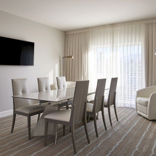 A modern dining room with a table and six chairs, a flat-screen TV on the wall, and a cozy armchair by sheer curtains.