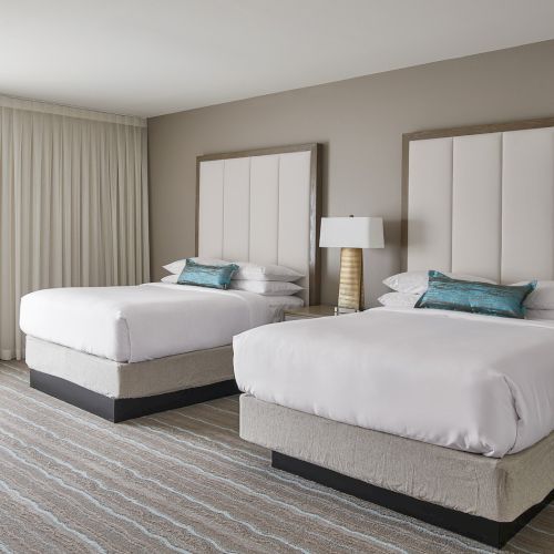 A hotel room with two double beds, decorative pillows, a lamp on a side table, and curtains drawn to the side on the left.