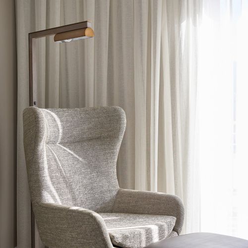 A cozy room corner featuring a gray armchair, footstool, floor lamp, and sheer curtains softly illuminated by natural light.