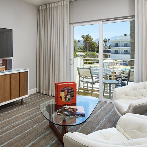 A modern living room with a TV, vinyl player, two chairs, a coffee table, lamp, and sliding doors opening to a balcony with outdoor seating.