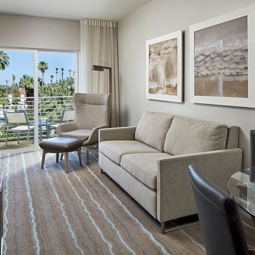 A modern hotel room with a sofa, chairs, a desk, and a large window overlooking palm trees, featuring neutral decor and wall art.