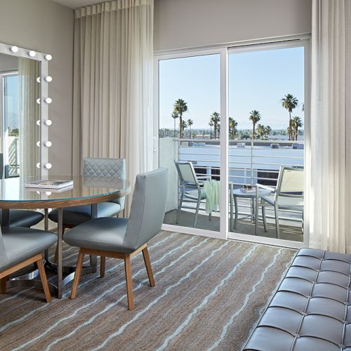 A modern room with a table, chairs, and a vanity mirror. There's a bed and sliding doors opening to a balcony with a view of palm trees.