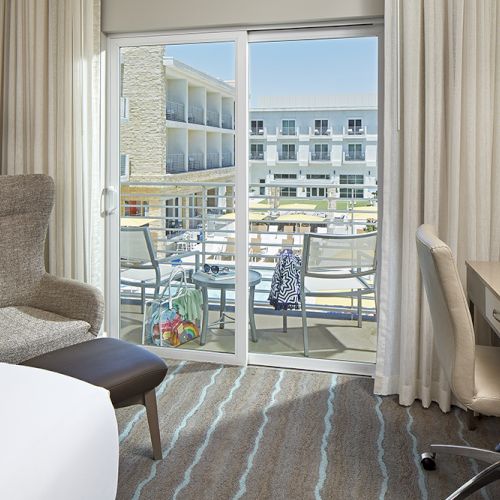 A hotel room with a chair, desk, and sliding doors leading to a balcony with a view of another building.