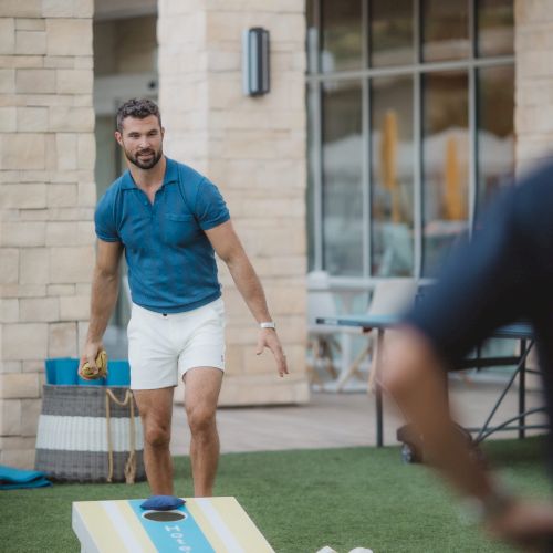 A person is playing cornhole outdoors, preparing to throw a bean bag towards the board on the grass.