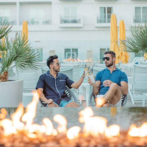 Two people are sitting outdoors, toasting drinks near a fire pit, surrounded by potted plants and patio furniture.