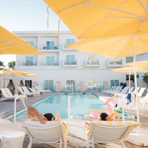 A sunny poolside scene with yellow umbrellas, lounge chairs, and people relaxing. Inflatable toys float in the pool.