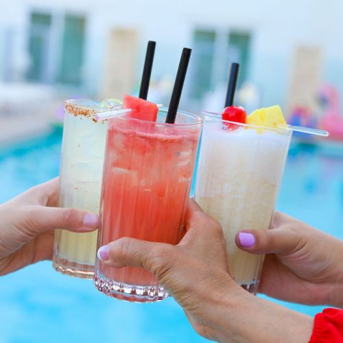 Three hands holding colorful cocktails with fruit garnishes by a pool. Bright, refreshing drinks for a relaxing summer day.