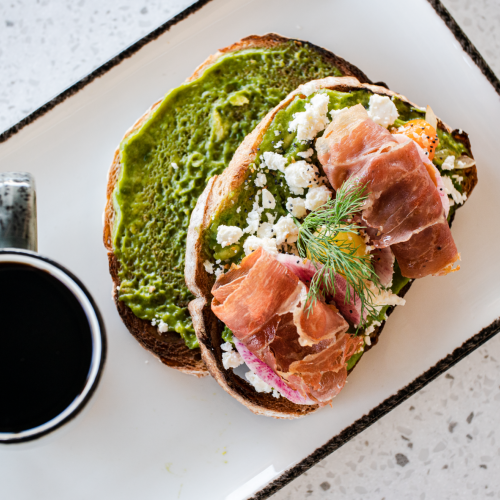 A plate with toast topped with green spread, cheese, and prosciutto next to a cup of black coffee on a speckled surface.