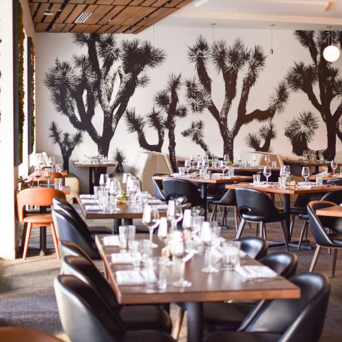 The image shows a stylish restaurant interior with tables, chairs, and Joshua tree murals on the wall. Modern lighting adds to the ambiance.