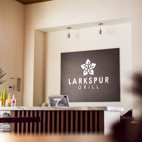 The image shows a reception area with a "Larkspur Grill" sign, a desk, plants, and natural light from the windows.