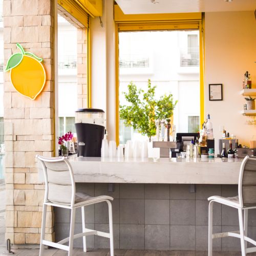 It's an outdoor bar with two chairs, a lemon sign, and a drink station in a sunny setting.