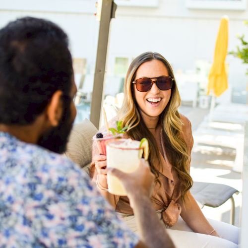 Two people are sitting by a pool, enjoying drinks, and smiling at each other.