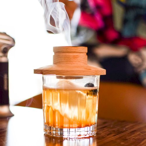 A cocktail glass with a smoky effect under a wooden lid, set on a wooden table with a colorful, blurred background.