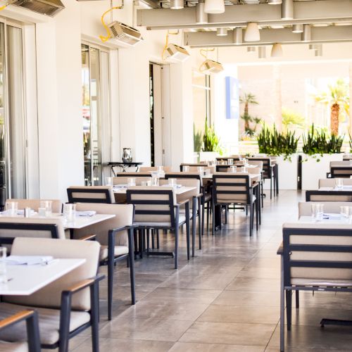 A bright, modern restaurant interior with tables and chairs arranged neatly, glass partitions, and potted plants in the background.