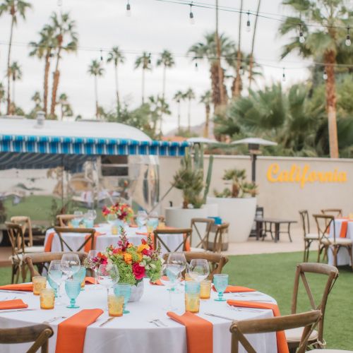 Outdoor event setup with tables, orange accents, flowers, and a vintage trailer. Surrounded by palm trees, furniture, and a California sign.