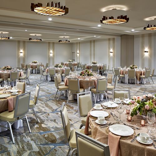 A banquet hall is elegantly set up with round tables, floral centerpieces, plates, and cups. Neutral tones and modern lighting complete the decor.