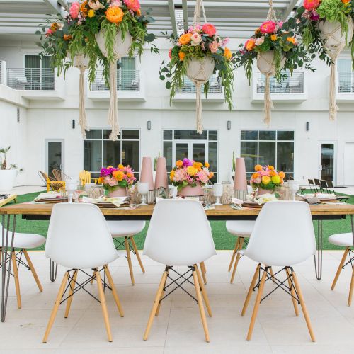 An outdoor dining setup with white chairs, colorful floral arrangements, and a modern table under a pergola adorned with hanging plants.