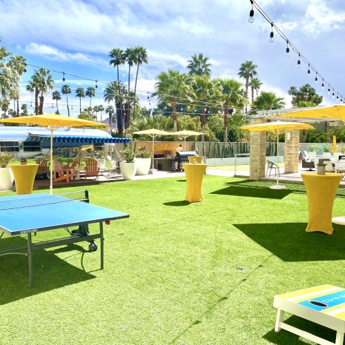 An outdoor space with a ping pong table, yellow umbrellas, palm trees, and a clear blue sky.