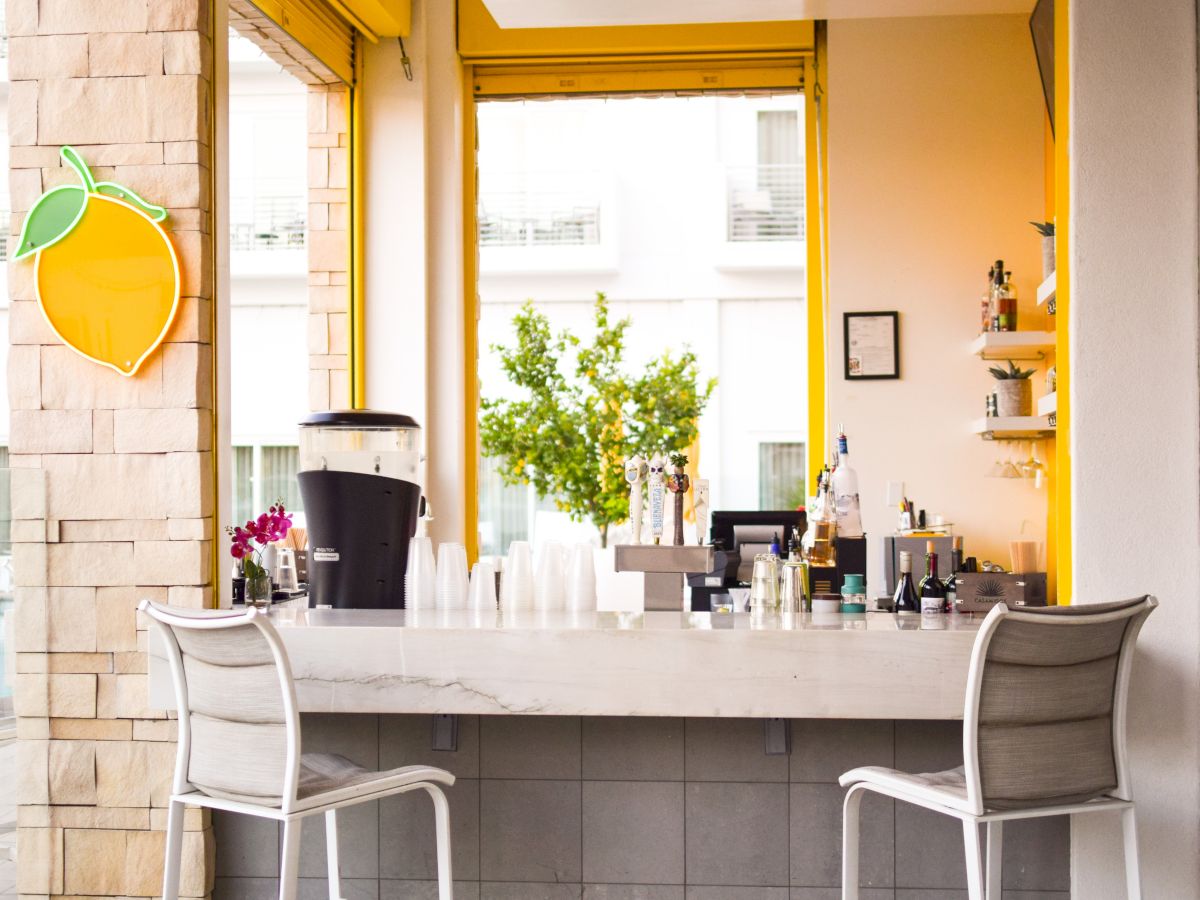 A bright bar with yellow accents, a lemon neon sign, seating, beverage dispensers, and bottles on shelves in a modern setting.