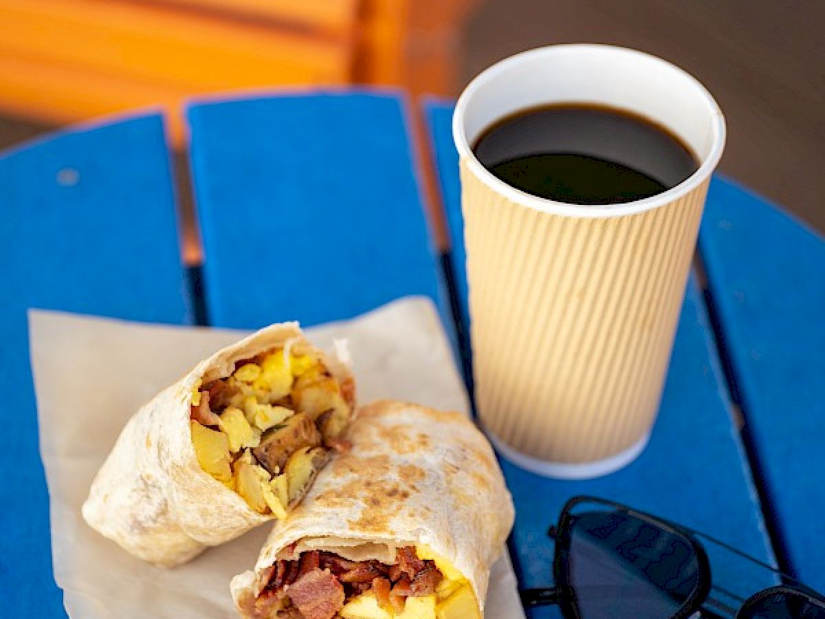 A breakfast burrito cut in half on a napkin, a cup of black coffee, and sunglasses on a blue table.